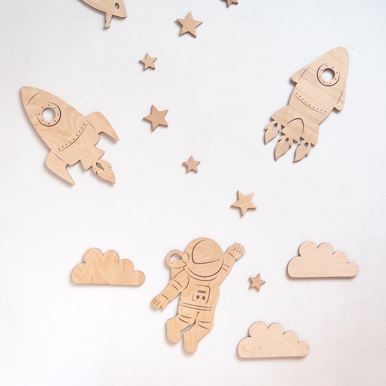 May include: Wooden astronaut, rocket, and cloud shapes in a space theme. The astronaut is in a space suit with a helmet and is floating in the air. The rocket is pointing upwards with flames coming out of the bottom. The clouds are white and fluffy.