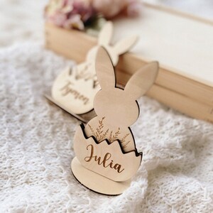 May include: Wooden Easter bunny place card with a name tag that says "Julia". The bunny is holding an egg with a floral design.