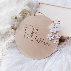 Peut inclure: Une pancarte ronde en bois avec le nom "Olivia" grav&eacute; en &eacute;criture cursive. La pancarte est d&eacute;cor&eacute;e de fleurs de marguerite blanches et poss&egrave;de un cordon en cuir brun pour l'accrocher.