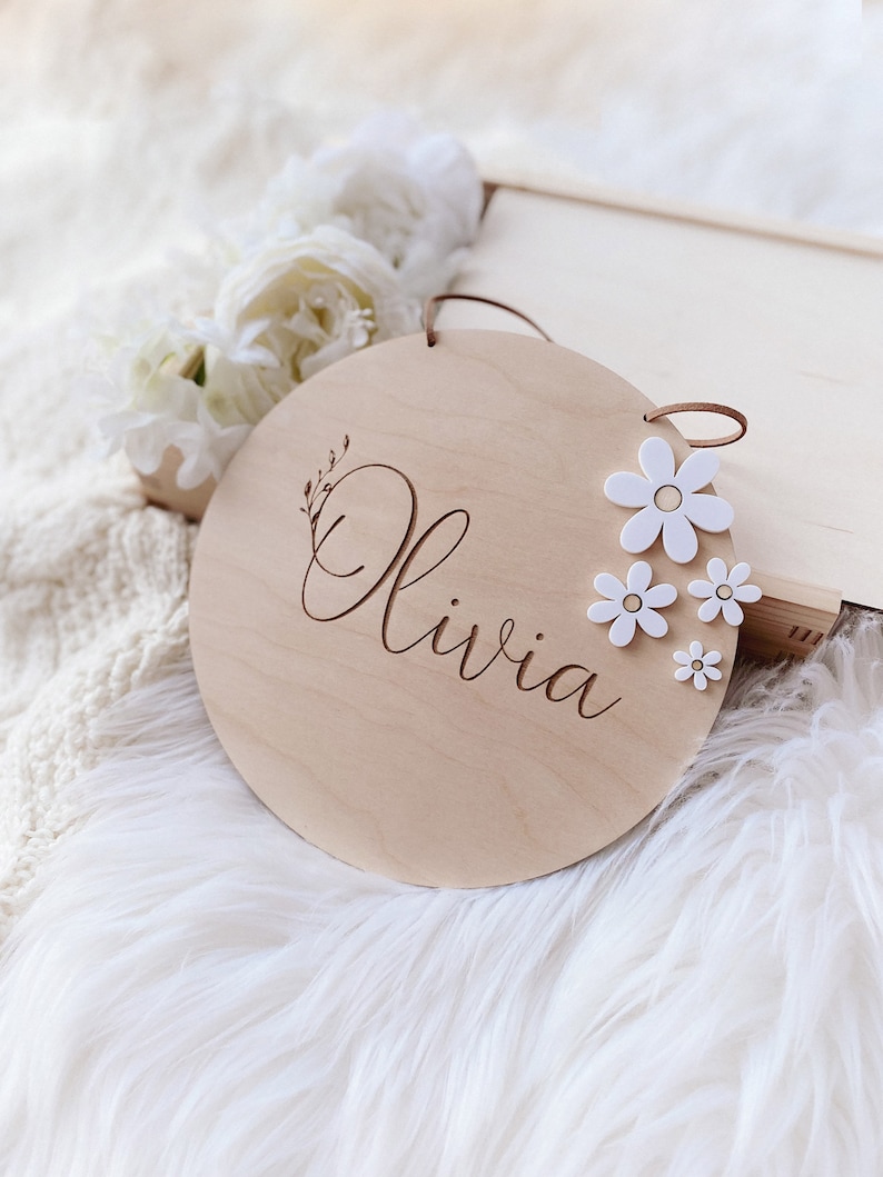 Peut inclure: Une pancarte ronde en bois avec le nom "Olivia" grav&eacute; en &eacute;criture cursive. La pancarte est d&eacute;cor&eacute;e de fleurs de marguerite blanches et poss&egrave;de un cordon en cuir brun pour l'accrocher.