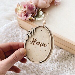 Peut inclure: Une d&eacute;coration en bois en forme d'&oelig;uf avec un motif de feuille et le nom "Henio" grav&eacute; dessus. La d&eacute;coration est suspendue &agrave; une ficelle et a une petite perle en bois en haut.