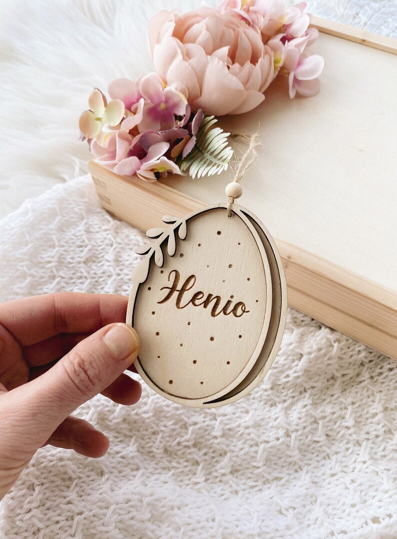 Peut inclure: Une d&eacute;coration en bois en forme d'&oelig;uf avec un motif de feuille et le nom "Henio" grav&eacute; dessus. La d&eacute;coration est suspendue &agrave; une ficelle et a une petite perle en bois en haut.