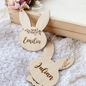 Op de afbeelding: Twee houten bunny-vormige ornamenten met de namen "Emilia" en "Julian" erop gegraveerd. De ornamenten zijn versierd met bloemmotieven en hebben een rustieke, natuurlijke uitstraling.
