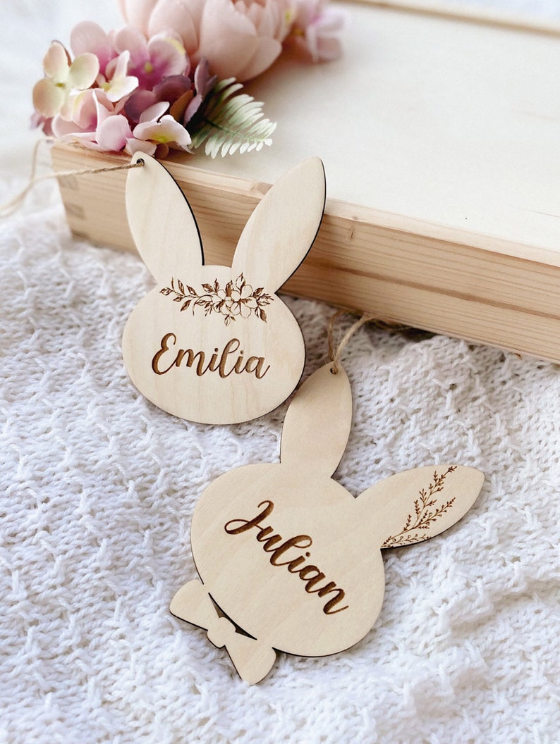 Op de afbeelding: Twee houten bunny-vormige ornamenten met de namen "Emilia" en "Julian" erop gegraveerd. De ornamenten zijn versierd met bloemmotieven en hebben een rustieke, natuurlijke uitstraling.
