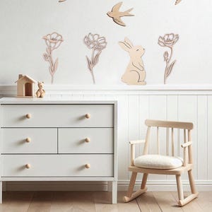 Puede incluir: Una escena de guarder&iacute;a con decoraciones de pared de madera de p&aacute;jaros, flores y un conejito. Una c&oacute;moda blanca con pomos de madera y una mecedora de madera est&aacute;n en primer plano. La habitaci&oacute;n tiene suelo de madera clara.
