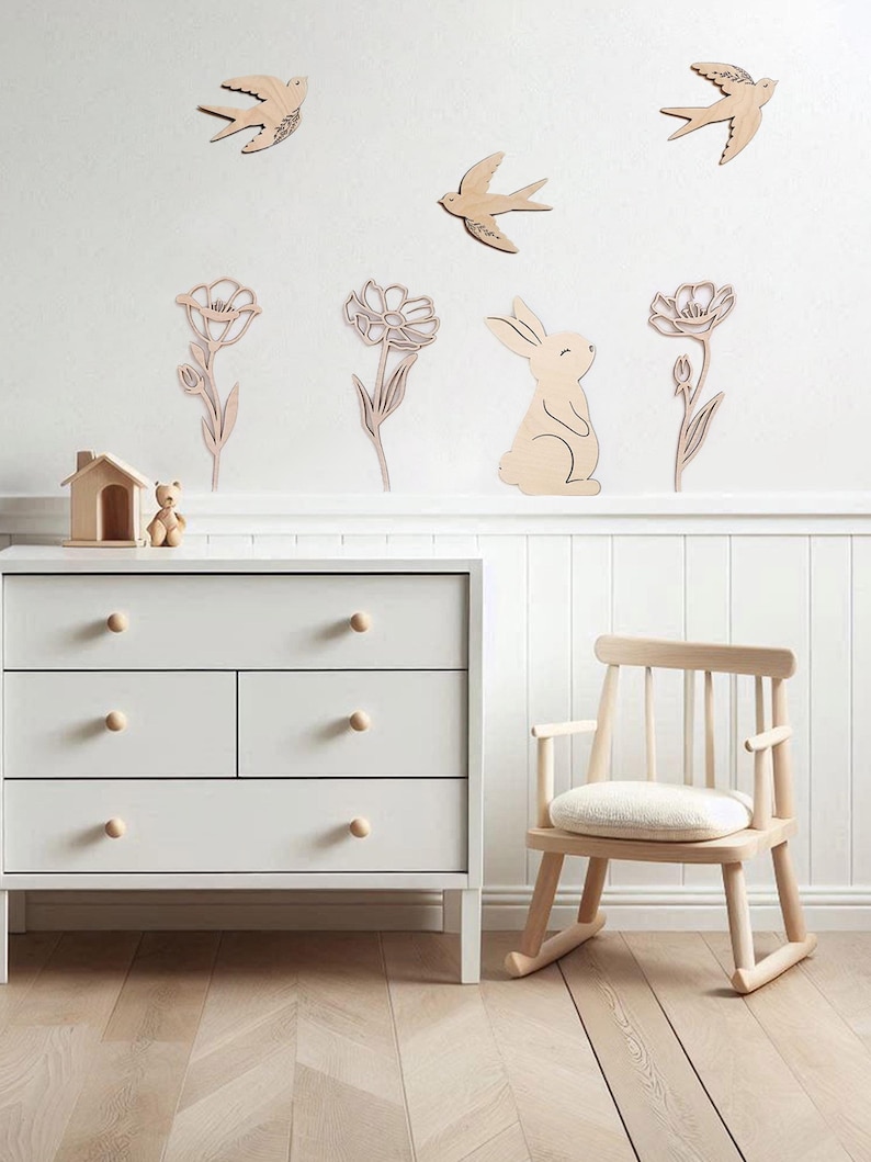 Puede incluir: Una escena de guarder&iacute;a con decoraciones de pared de madera de p&aacute;jaros, flores y un conejito. Una c&oacute;moda blanca con pomos de madera y una mecedora de madera est&aacute;n en primer plano. La habitaci&oacute;n tiene suelo de madera clara.
