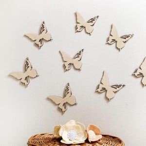 Op de afbeelding: Negen houten vlinderuitsparingen in verschillende maten zijn op een witte muur gerangschikt. De vlinders zijn lichtbeige van kleur met ingewikkelde vleugeld details. Hieronder staat een geweven mand met viltbloemen in crème en perzik.