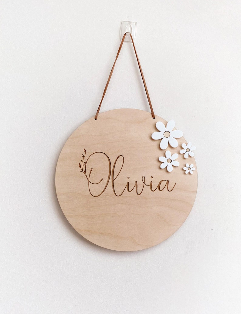 Peut inclure: Une pancarte ronde en bois avec le nom "Olivia" grav&eacute; en &eacute;criture cursive. La pancarte est d&eacute;cor&eacute;e de fleurs en bois blanc et poss&egrave;de une sangle de suspension en cuir marron.