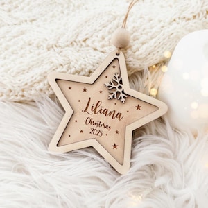 Puede incluir: Adorno navideño de madera en forma de estrella con el nombre "Liliana" y el año "2025" grabados. El adorno presenta detalles de copos de nieve y estrellas. Cuelga de un cordón natural y una cuenta de madera.
