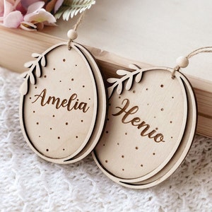 Peut inclure: Deux d&eacute;corations d'&oelig;ufs de P&acirc;ques en bois avec des crochets en ficelle. Les &oelig;ufs sont d&eacute;cor&eacute;s de pois blancs et d'une branche feuillue. Les noms Amelia et Henio sont grav&eacute;s sur les &oelig;ufs.