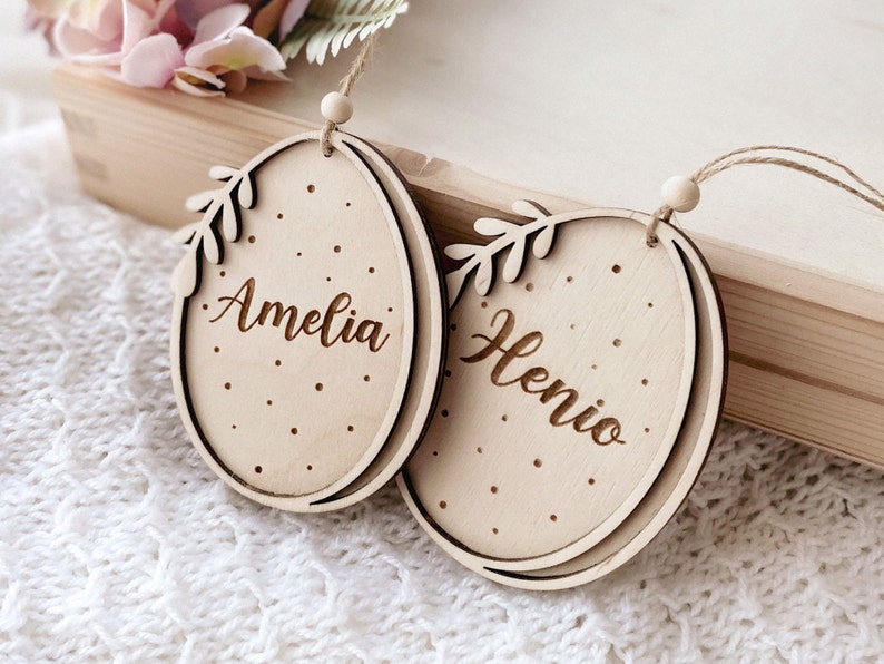 Peut inclure: Deux d&eacute;corations d'&oelig;ufs de P&acirc;ques en bois avec des crochets en ficelle. Les &oelig;ufs sont d&eacute;cor&eacute;s de pois blancs et d'une branche feuillue. Les noms Amelia et Henio sont grav&eacute;s sur les &oelig;ufs.
