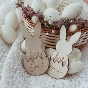 May include: Two wooden bunny-shaped Easter egg holders with the names "Emil" and "Hania" engraved on them. The holders are surrounded by white speckled Easter eggs and a wicker basket filled with white eggs and purple flowers.