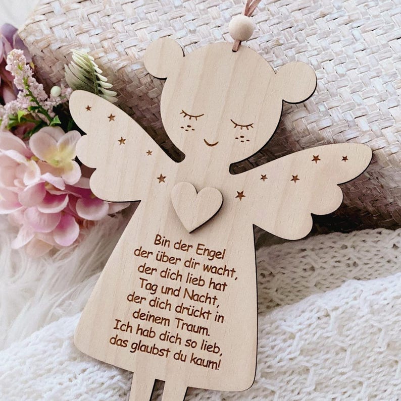 Puede incluir: Adorno de &aacute;ngel de madera con detalles de coraz&oacute;n y estrellas. El &aacute;ngel tiene una cabeza de oso y texto en alem&aacute;n. El adorno cuelga de una cuerda.