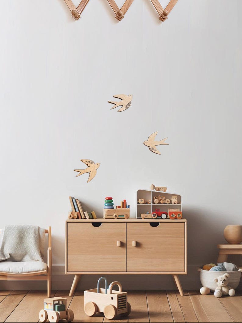 Puede incluir: Un mueble de madera clara con dos puertas y un conjunto de juguetes de madera. Tres golondrinas de madera est&aacute;n montadas en la pared blanca sobre el mueble. Una silla de madera con una manta y un coche de juguete est&aacute;n en primer plano.