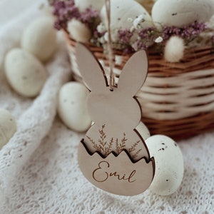 May include: A wooden Easter bunny ornament with a cutout egg shape and the name "Emil" engraved on it. The ornament is hanging from a string and is surrounded by white eggs in a wicker basket.
