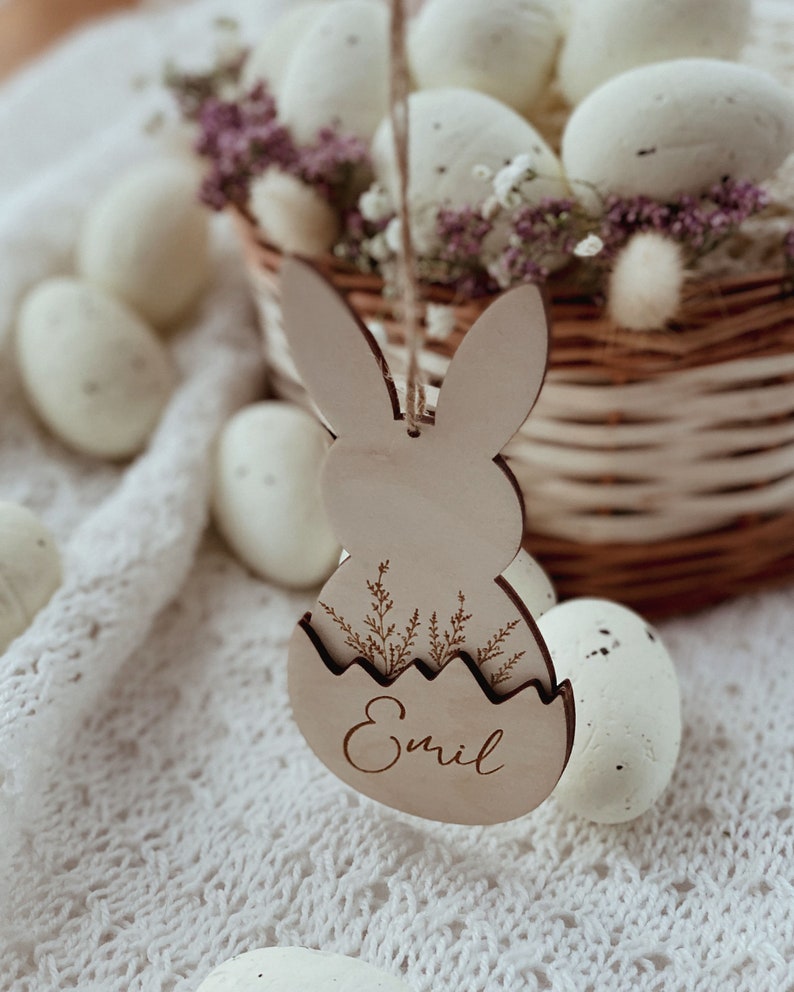 May include: A wooden Easter bunny ornament with a cutout egg shape and the name "Emil" engraved on it. The ornament is hanging from a string and is surrounded by white eggs in a wicker basket.