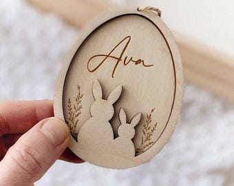 Op de afbeelding: Houten paasei-ornament met de naam "Ava" in cursief schrift. Het ei heeft een uitgesneden ontwerp van twee konijnen en takjes. Er is een touwlus bevestigd om op te hangen.