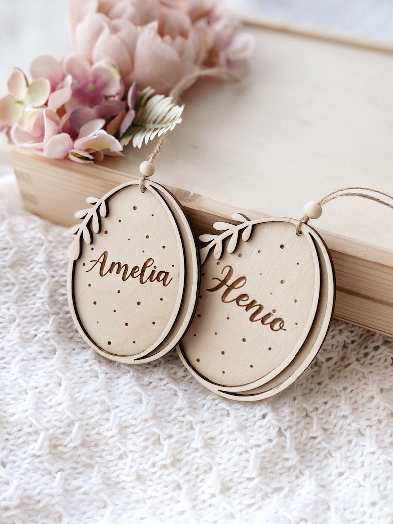 Peut inclure: Deux d&eacute;corations d'&oelig;ufs de P&acirc;ques en bois avec les noms Amelia et Henio grav&eacute;s dessus. Les &oelig;ufs sont d&eacute;cor&eacute;s de petits points blancs et ont un motif de feuilles.