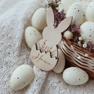 May include: A wooden bunny-shaped Easter egg holder with the name "Hania" engraved on it. The holder is filled with white speckled Easter eggs and surrounded by white flowers in a wicker basket.