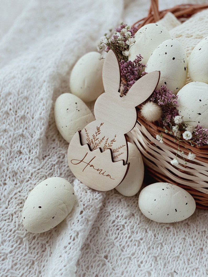 May include: A wooden bunny-shaped Easter egg holder with the name "Hania" engraved on it. The holder is filled with white speckled Easter eggs and surrounded by white flowers in a wicker basket.