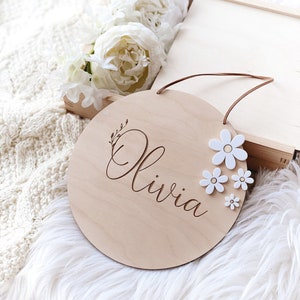 Peut inclure: Une pancarte ronde en bois avec le nom "Olivia" grav&eacute; en &eacute;criture cursive. La pancarte est d&eacute;cor&eacute;e de fleurs blanches et poss&egrave;de un cordon brun pour l'accrocher.