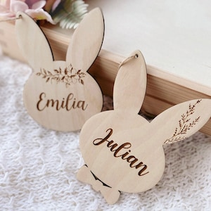 Op de afbeelding: Twee houten bunny-vormige ornamenten met de namen "Emilia" en "Julian" erop gegraveerd. Elk ornament heeft een bloemmotief en een klein strikje.