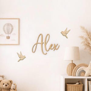 Op de afbeelding: Een lichtgekleurde houten wanddecoratie met de naam Alex in een cursief lettertype, vergezeld door twee kleine houten vogel-silhouetten aan weerszijden van de naam, wat een decoratieve opstelling vormt voor op een muur.