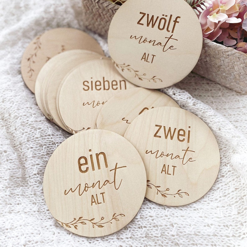 May include: A set of twelve wooden baby milestone discs with a floral design and the text "ein monat alt", "zwei monate alt", "sieben monate alt", and "zw&ouml;lf monate alt".