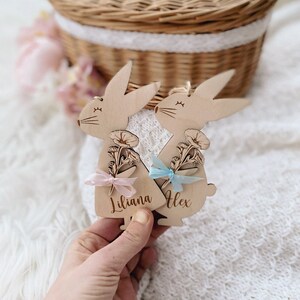 May include: Two wooden bunny ornaments with floral designs and personalised names, Liliana and Alex, with pink and blue ribbon accents.