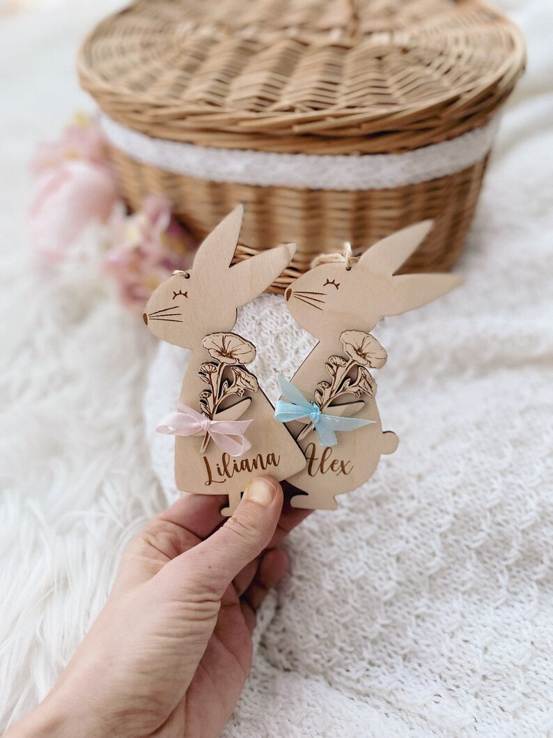May include: Two wooden bunny ornaments with floral designs and personalised names, Liliana and Alex, with pink and blue ribbon accents.