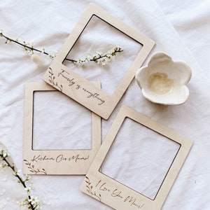 May include: Three wooden Polaroid-style photo frames, each with a message: "Family is everything," "I Love You Mom!," and "Kocham Cię Mamo!" Floral details and a small white flower-shaped dish are also present. The frames are light-coloured.