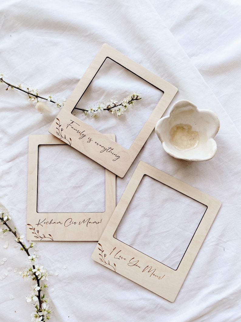 May include: Three wooden Polaroid-style photo frames, each with a message: "Family is everything," "I Love You Mom!," and "Kocham Cię Mamo!" Floral details and a small white flower-shaped dish are also present. The frames are light-coloured.