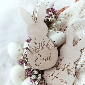 May include: Two wooden Easter bunny ornaments with the names "Emil" and "Hania" engraved on them. The ornaments are decorated with a floral design and are surrounded by white and pink flowers.