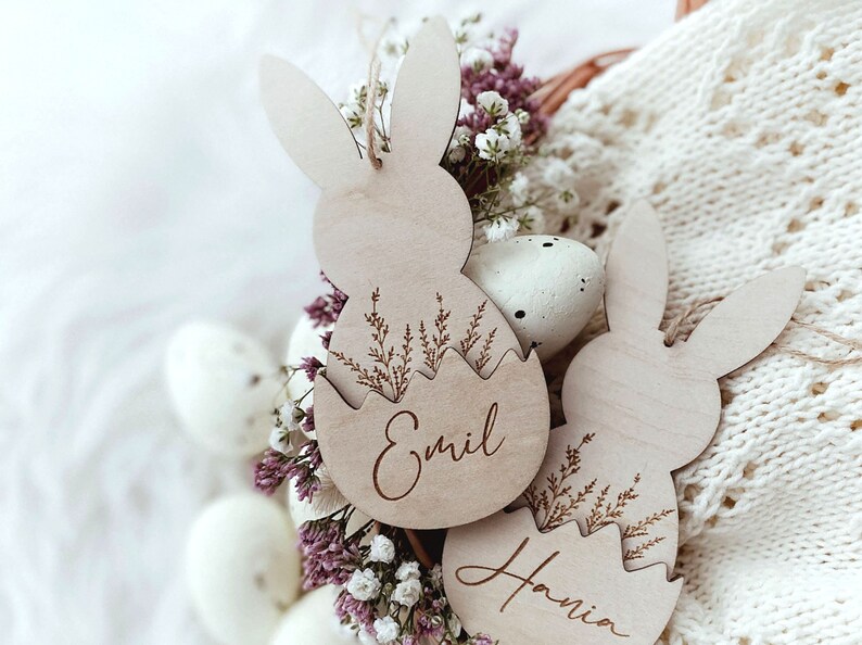 May include: Two wooden Easter bunny ornaments with the names "Emil" and "Hania" engraved on them. The ornaments are decorated with a floral design and are surrounded by white and pink flowers.