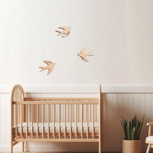 Puede incluir: Una cuna de madera clara con un colch&oacute;n, un juego de trenes de juguete de madera y tres decoraciones de pared de p&aacute;jaros de madera. La habitaci&oacute;n tiene una paleta de colores neutros con una planta en una cesta tejida y un perchero de madera.