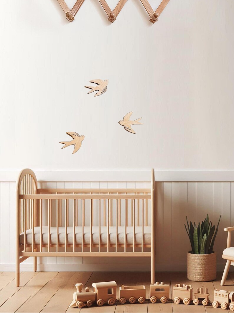 Puede incluir: Una cuna de madera clara con un colch&oacute;n, un juego de trenes de juguete de madera y tres decoraciones de pared de p&aacute;jaros de madera. La habitaci&oacute;n tiene una paleta de colores neutros con una planta en una cesta tejida y un perchero de madera.