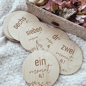 May include: Wooden baby milestone cards with the words "ein monat alt", "zwei monate alt", "drei monate alt", "sieben monate alt", and "sechs monate alt" written on them. The cards are arranged in a circle on a white knitted blanket.