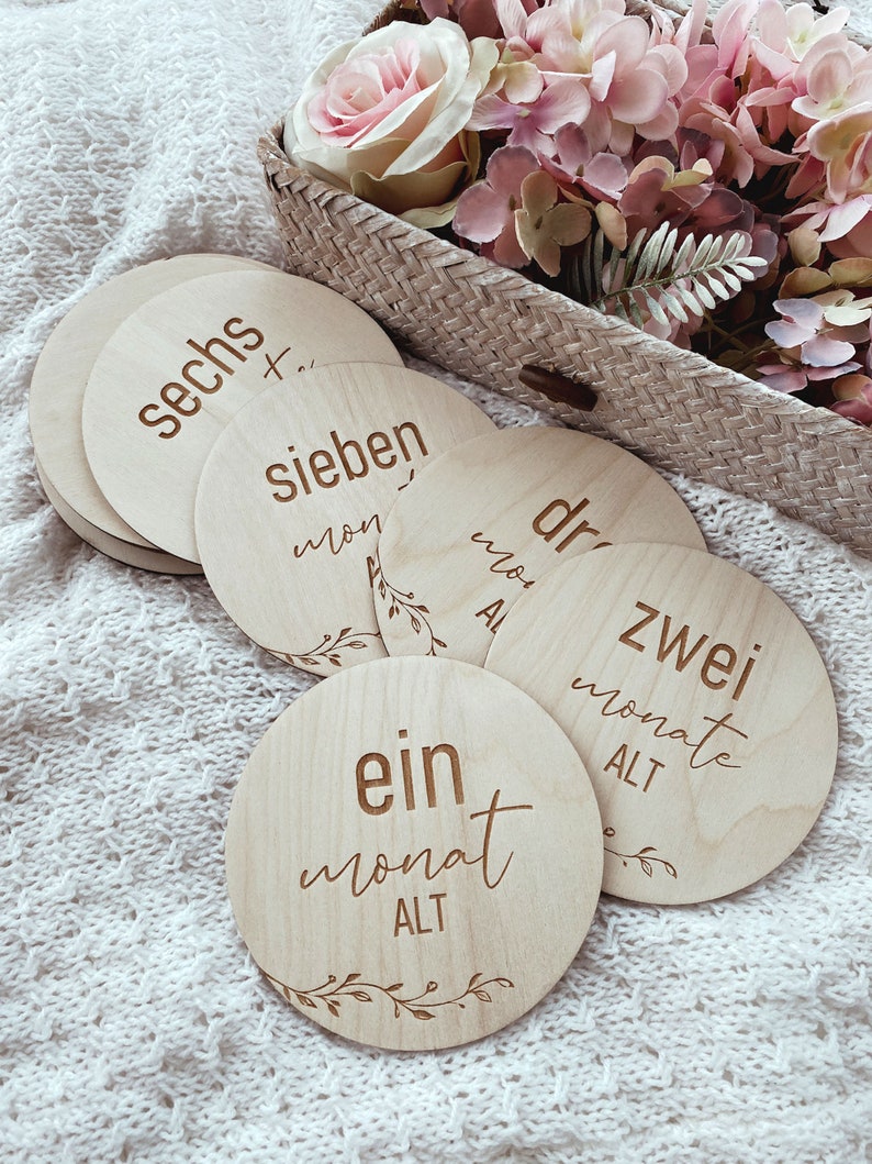 May include: Wooden baby milestone cards with the words "ein monat alt", "zwei monate alt", "drei monate alt", "sieben monate alt", and "sechs monate alt" written on them. The cards are arranged in a circle on a white knitted blanket.