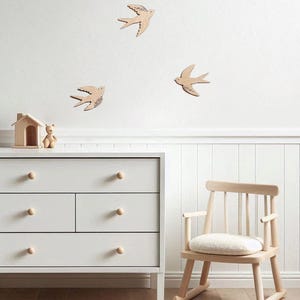 Puede incluir: Escena de guarder&iacute;a con decoraci&oacute;n de pared de p&aacute;jaros de madera, una c&oacute;moda blanca con pomos de madera, una mecedora de madera y una peque&ntilde;a casa de madera con un oso de peluche. La habitaci&oacute;n tiene un ambiente luminoso.