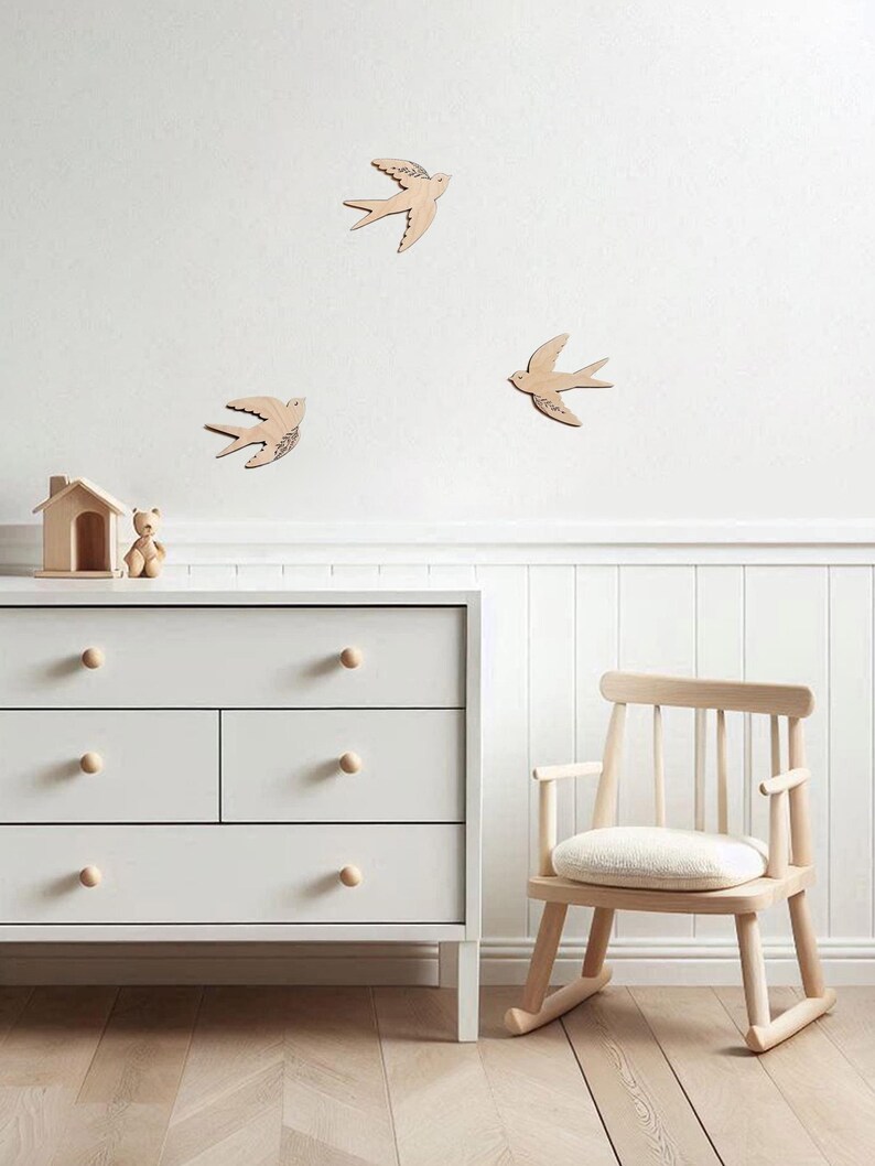 Puede incluir: Escena de guarder&iacute;a con decoraci&oacute;n de pared de p&aacute;jaros de madera, una c&oacute;moda blanca con pomos de madera, una mecedora de madera y una peque&ntilde;a casa de madera con un oso de peluche. La habitaci&oacute;n tiene un ambiente luminoso.