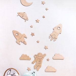 May include: Wooden wall decor featuring a rocket, astronaut, UFO, clouds, moon, and stars. The decor is in a light brown color and is perfect for a child's room.