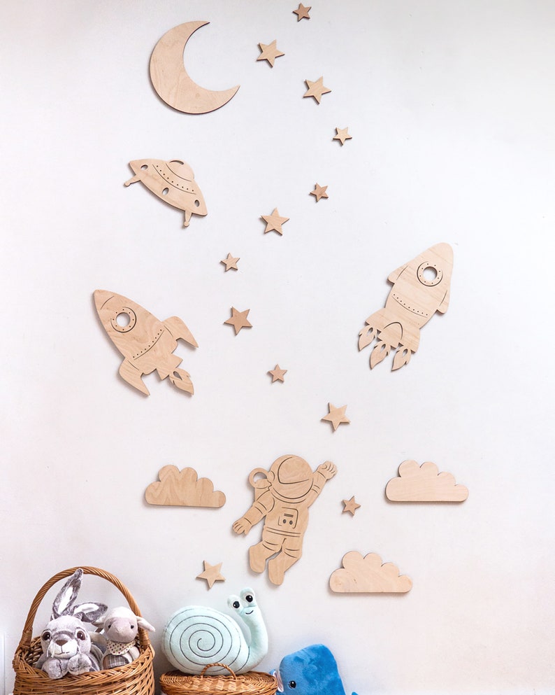 May include: Wooden wall decor featuring a rocket, astronaut, UFO, clouds, moon, and stars. The decor is in a light brown color and is perfect for a child's room.