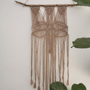 May include: A macrame wall hanging with a butterfly design, made with natural beige cord and a branch at the top. The hanging has long tassels at the bottom.
