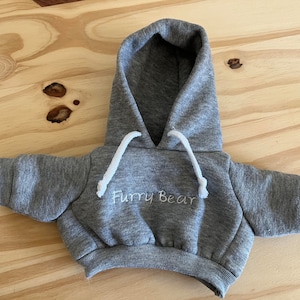 May include: A gray miniature hoodie with a hood and white drawstrings. The words "Furry Bear" are embroidered in white on the front. The hoodie is designed for a small stuffed animal or doll.