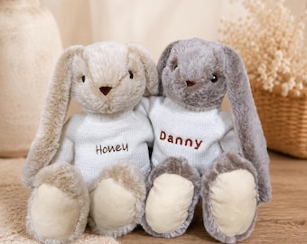 May include: Two plush stuffed bunnies with long ears, one beige and one gray, each wearing a white knitted sweater. The beige bunny has the name Honey embroidered in brown on its sweater, and the gray bunny has the name Danny embroidered in brown.