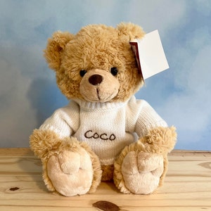 May include: A plush teddy bear with light brown fur, wearing a white sweater embroidered with the name "Coco". The bear has a brown nose and black eyes. A blank white tag hangs from its ear. The bear is sitting on a wooden surface.