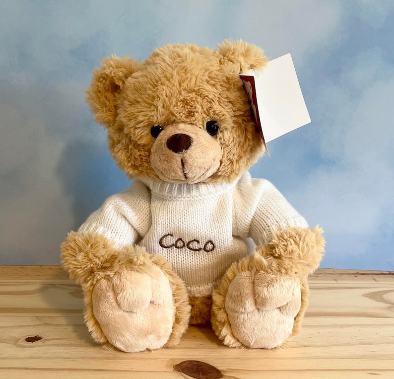 May include: A plush teddy bear with light brown fur, wearing a white sweater embroidered with the name "Coco". The bear has a brown nose and black eyes. A blank white tag hangs from its ear. The bear is sitting on a wooden surface.