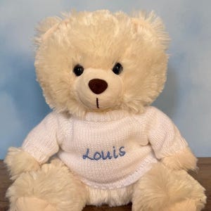 May include: A plush, cream-colored teddy bear with the text "Furry Bear" above it. The bear wears a white sweater with the name "Louis" embroidered in blue. The word "White" is below the bear.