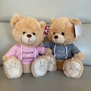 May include: Two plush teddy bears with tan fur. One bear wears a pink hoodie with "Princess Emma" embroidered on it, and the other wears a gray hoodie with "King Hector". Both have brown eyes and noses.