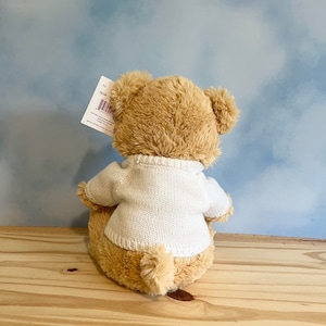 May include: A plush teddy bear, viewed from the back, wearing a white sweater. The bear has light brown fur and is sitting on a wooden surface. A tag is attached to the bear's ear. The background is a light blue sky.